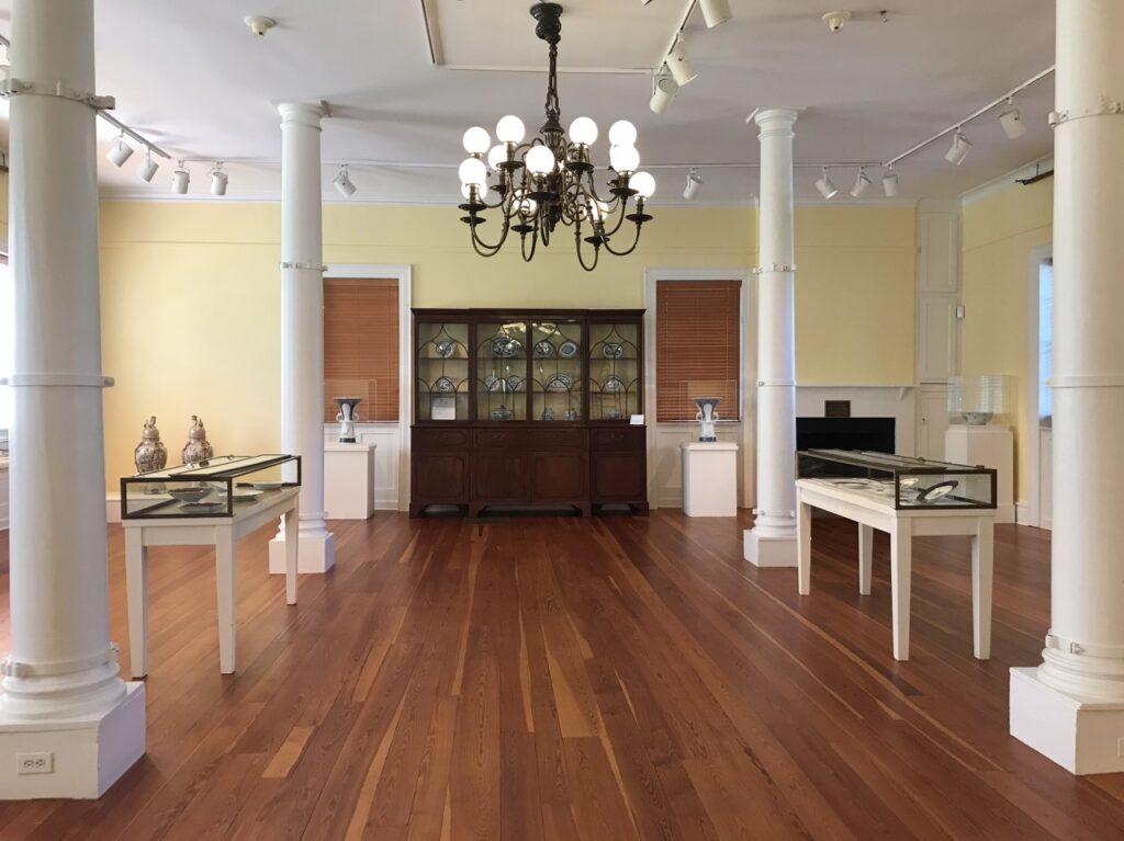 An open gallery has glass cases on either side of the room. A large chandelier hangs in the center. At the back of the room is a large wooden breakfront cabinet with china pieces inside.