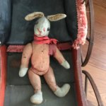An old slightly tattered stuffed rabbit toy rests inside of an antique stroller. The rabbit has a red ribbon tied in a bow around the neck.