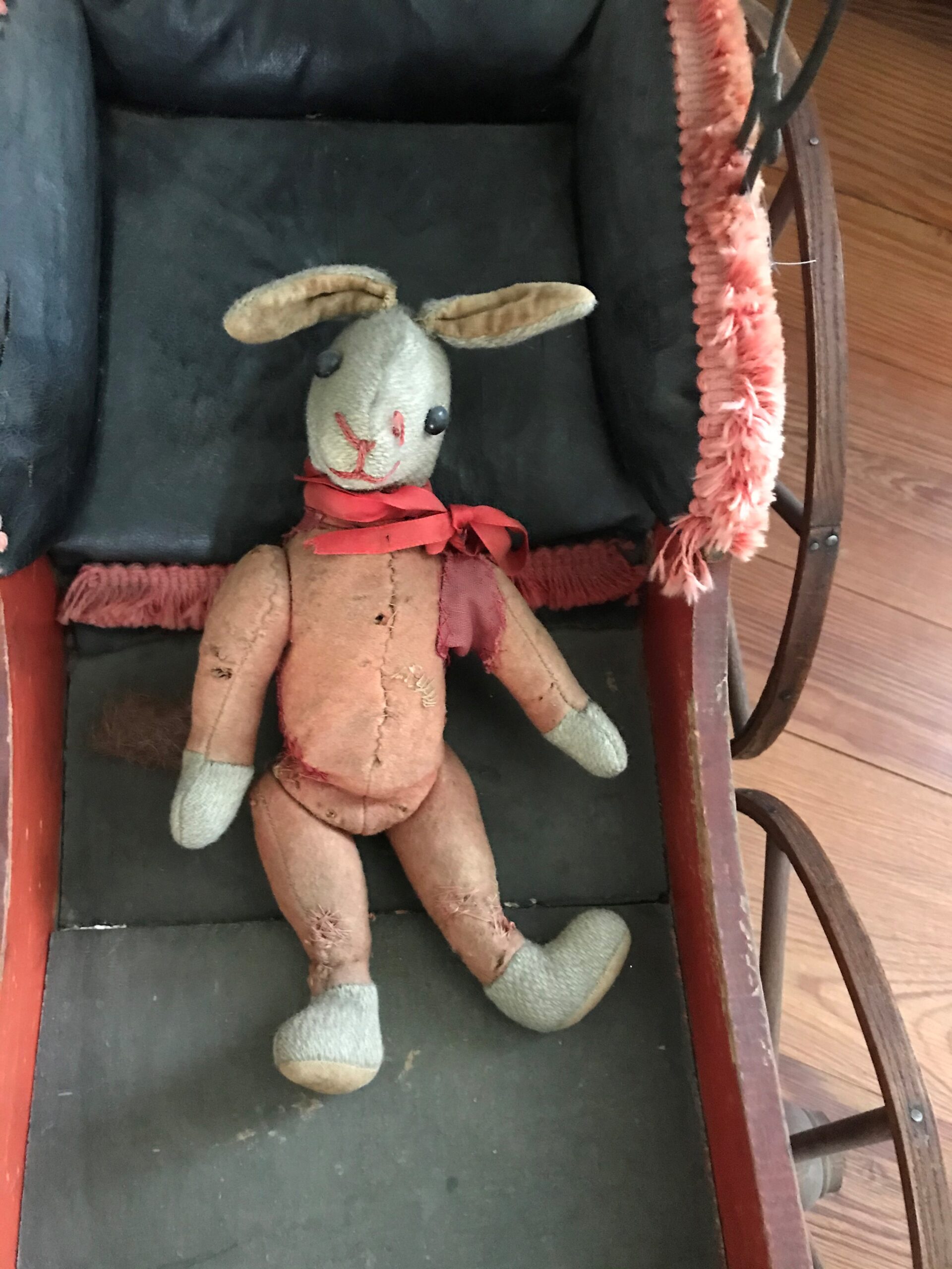 An old slightly tattered stuffed rabbit toy rests inside of an antique stroller. The rabbit has a red ribbon tied in a bow around the neck.