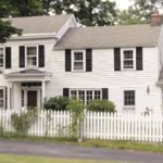 A white house with several windows and black shutters. A white picket fence is in front of the house.