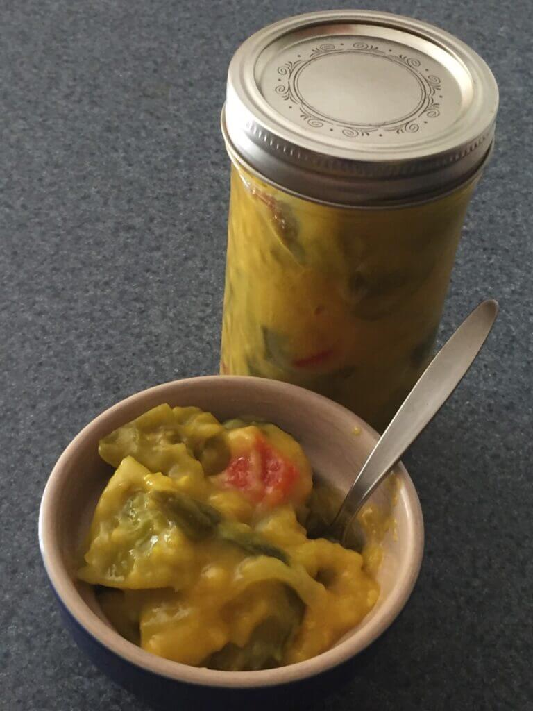 A glass jar of sweet mustard pickles sits next to a bowl of sweet mustard pickles.