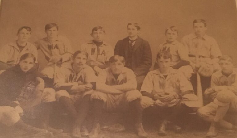 Seis hombres sentados en el fondo de la foto con cinco hombres sentados delante de ellos. Llevan uniformes de béisbol. Un hombre de la última fila lleva un traje.