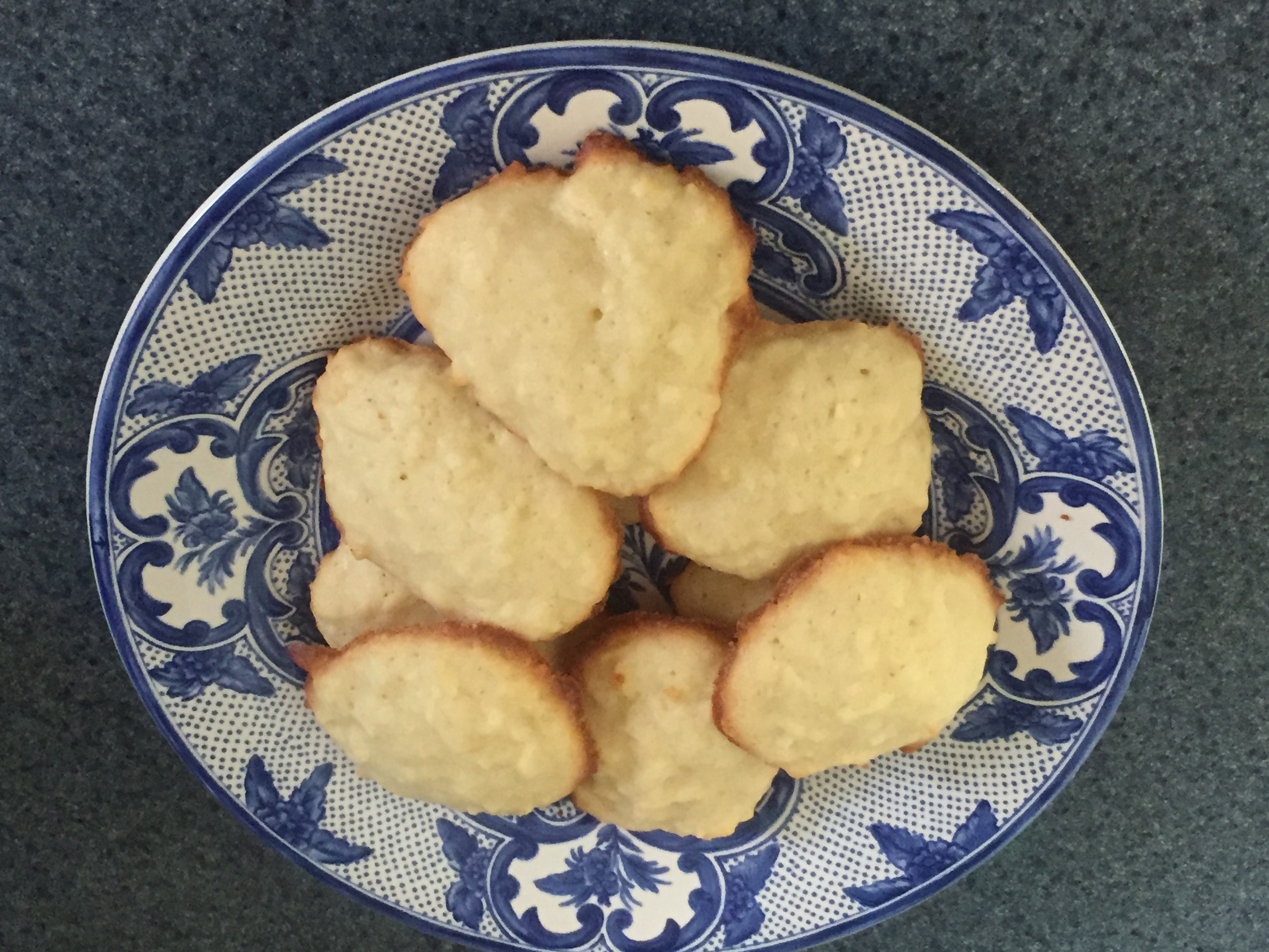 Eight lightly browned coconut jumbal cookies are stacked on a decorative blue and white porcelain plate.
