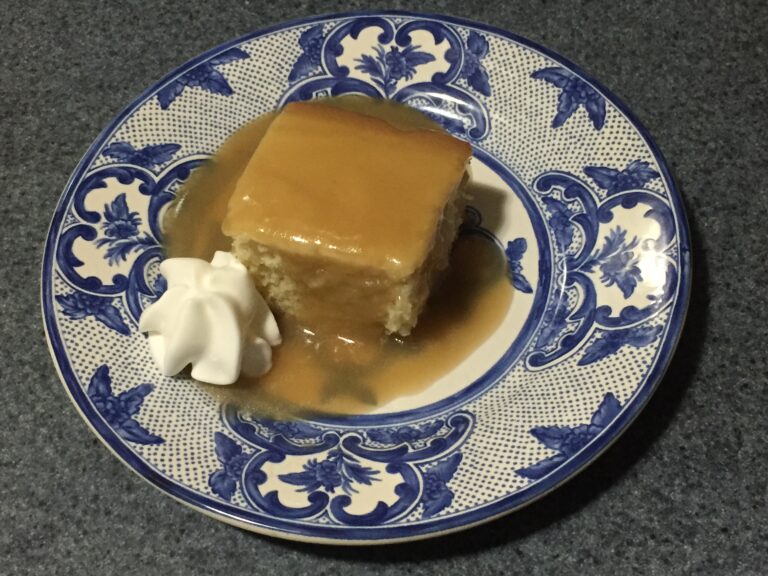 A baked slice of cottage pudding with sauce poured over top and whipped cream to the left.