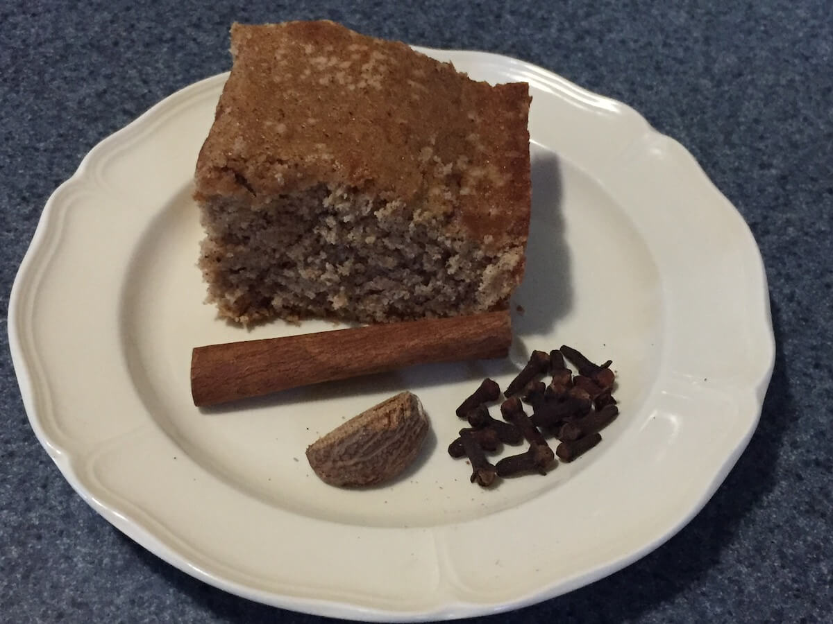 A baked slice of clove cake on a plate with whole cinnamon stick, ginger, and cloves.