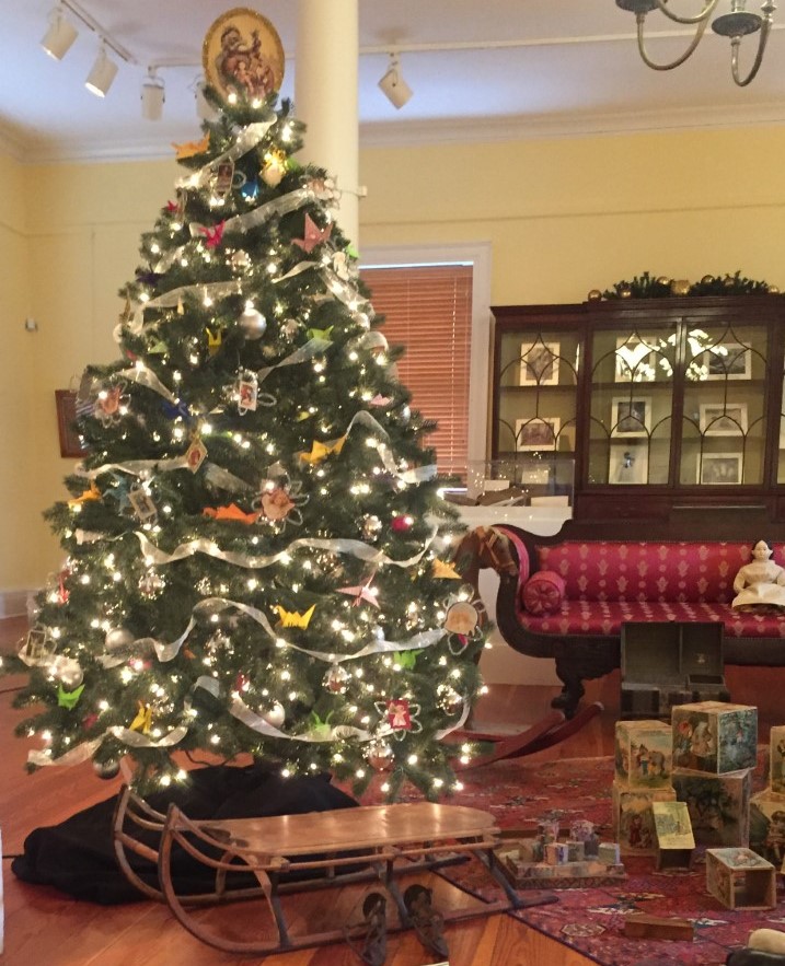 A Christmas tree decorated with paper ornaments and ribbon stands next to a collection of antique toys.