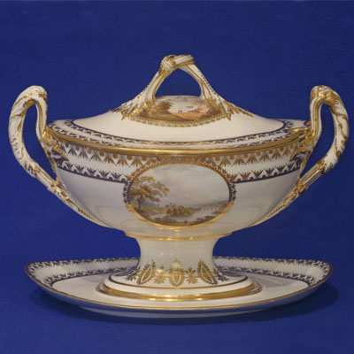 Ceramic tureen with decorative gold features and an image of trees and a body of water in the center. Handles on either side of the vessel and on the lid on top.