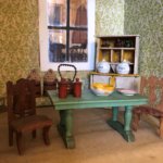 Up close shot of a dollhouse containing two chairs, a table with small bowl and plate on top, and shelf.