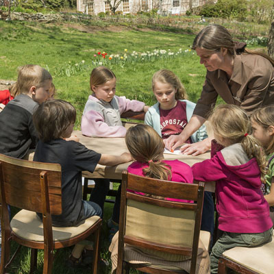 Several children sit around a table outdoors with a woman instructing.