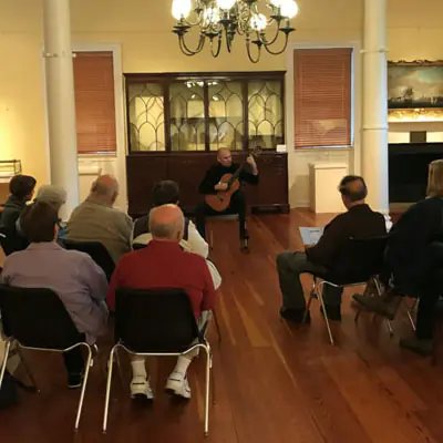 At the center, a sitting man plays guitar. Several people sit around watching him in a gallery.