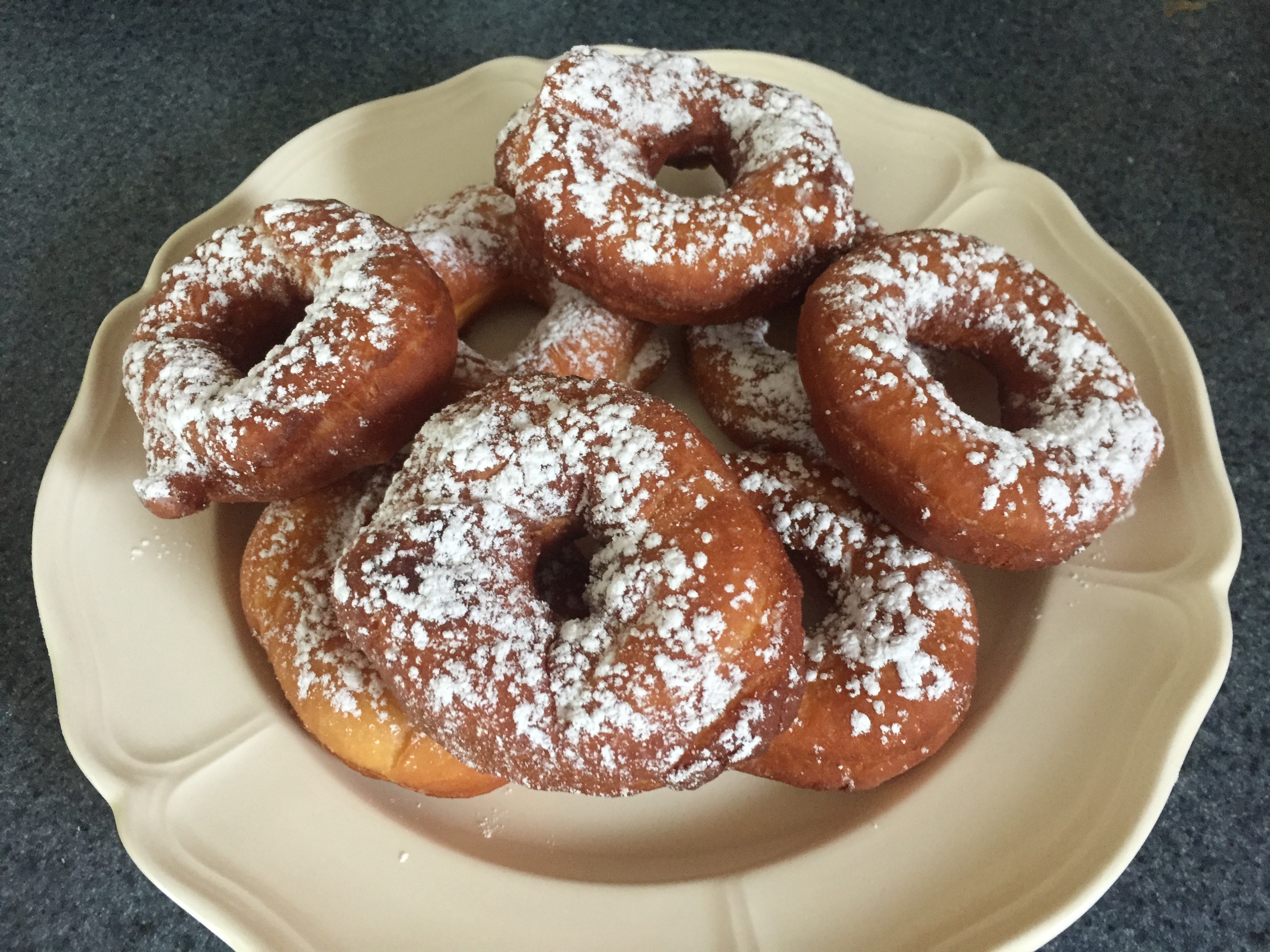 8 freshly baked donuts dusted with confectioner's sugar set on a white ceramic plate.