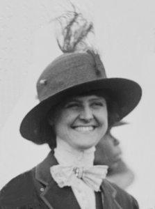 A black and white candid portrait photograph of a young, smiling woman. She is dressed in Victorian clothing and wears a wide-brimmed hat with a feather in it.