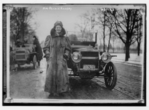 Black and white photograph of a young, smiling woman standing in front of an automobile that looks like a Model T Ford. It has a NJ license plate and she is wearing a full length driving coat.