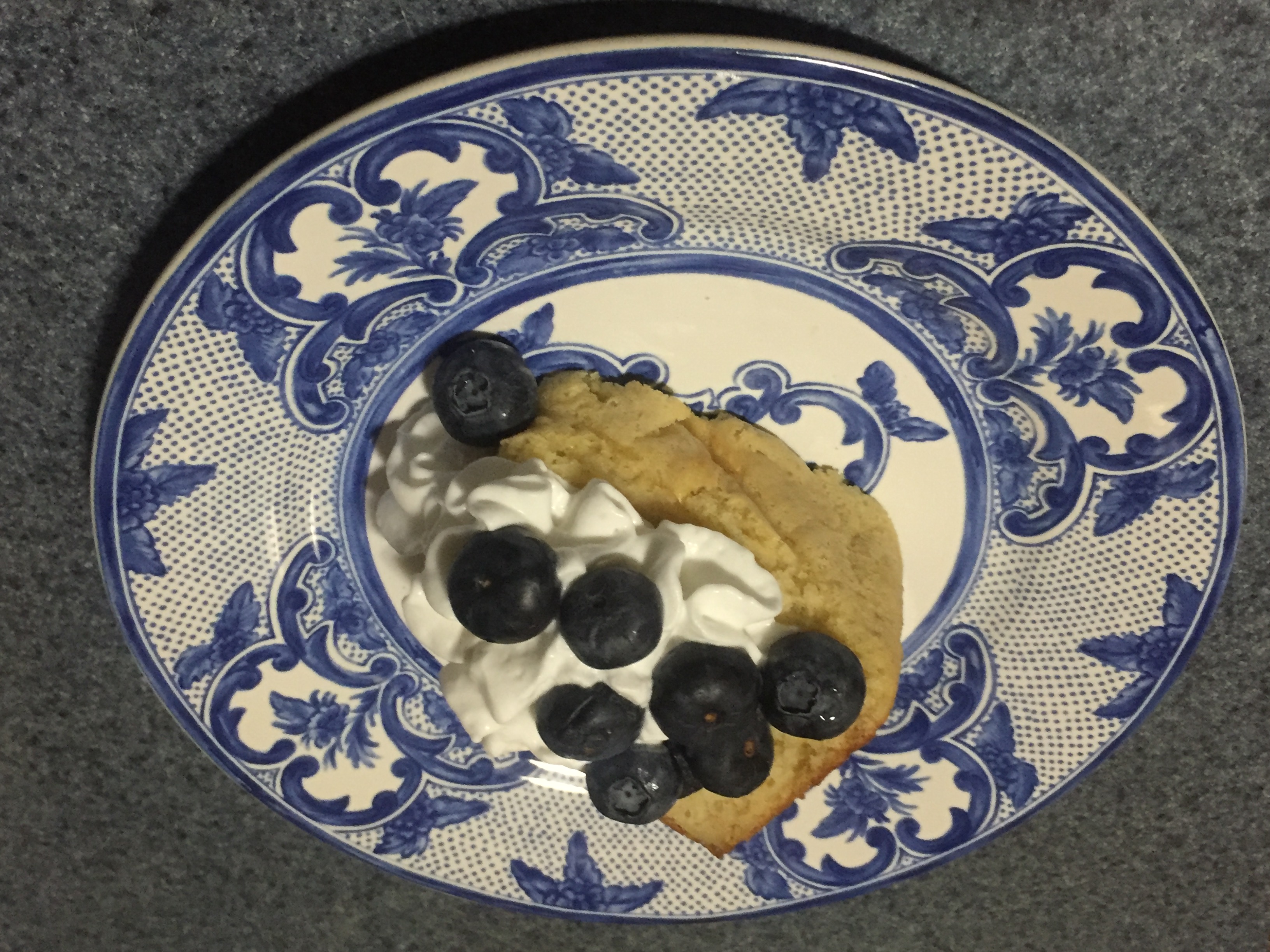 A slice of yellow sponge cake topped with whipped cream and blueberries on a blue and white china plate.