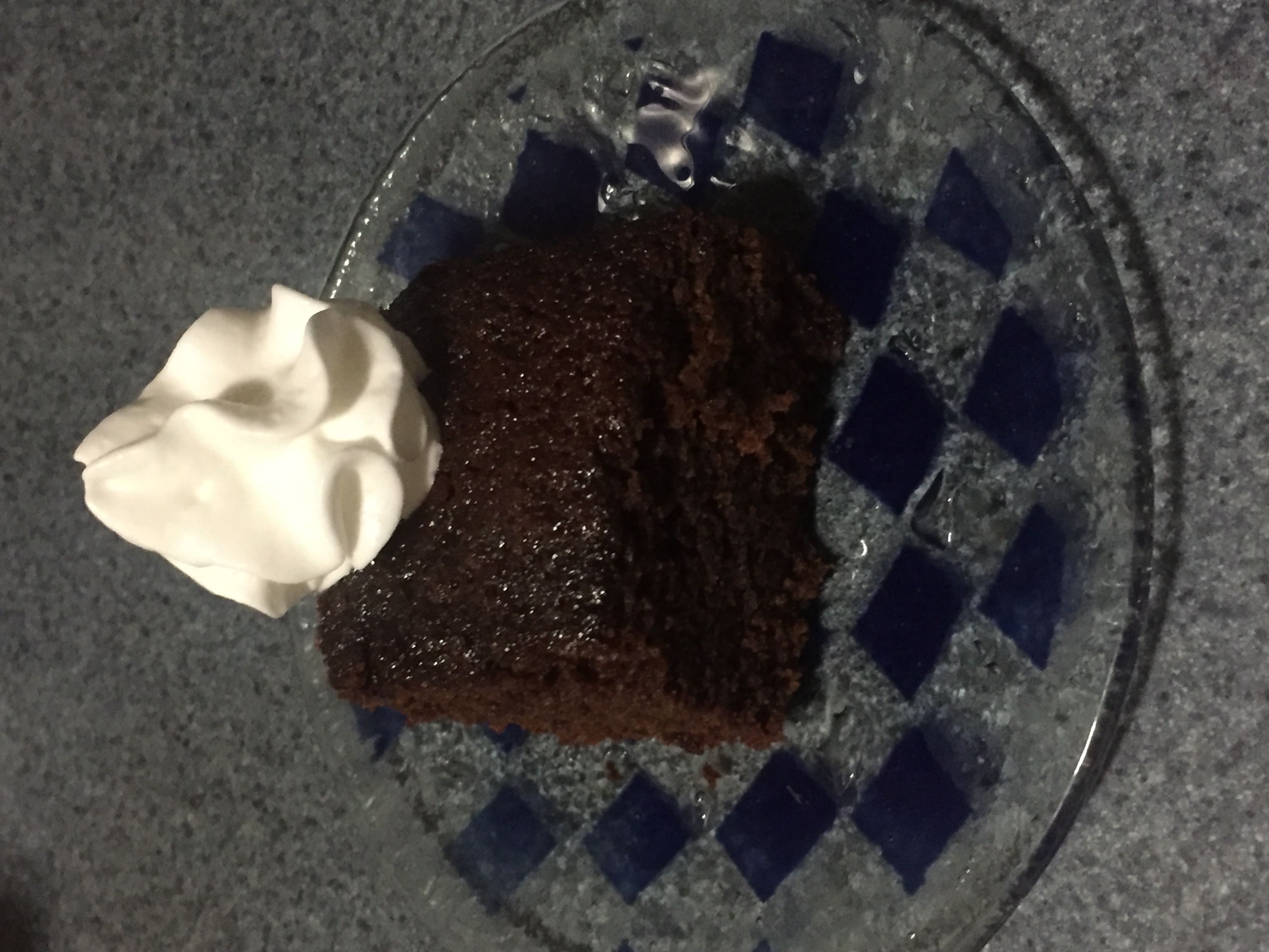Square slice of a dark molasses cake with whipped cream on top set on a blue-checked glass dish.