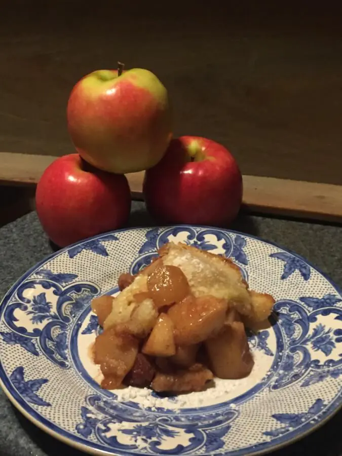 Un postre de crumble de manzana descansa sobre un plato decorativo. Detrás del plato hay tres manzanas rojas en forma de pirámide.
