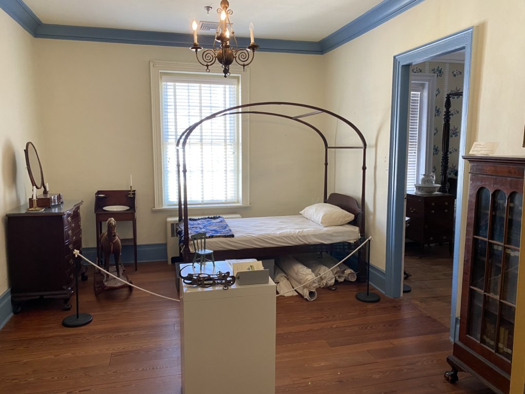 A bed with metal canopy frame sits in the corner of the room. To the left of the bed is a wooden washbasin, wooden rocking horse, and wooden armoire with mirror. In the center of the room is a pedestal with objects that can be touched. To the right, there is a wooden enclosed bookcase.