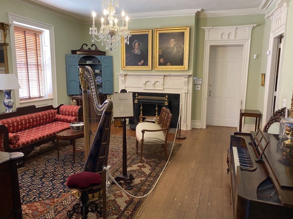To the left of the room sits a long red couch, tall wooden harp, music stand, and red stool. In the back corner is a blue cabinet with porcelain. Above the carved fireplace hangs the portraits of Louisa and George Macculloch. To the right sits a wooden piano.
