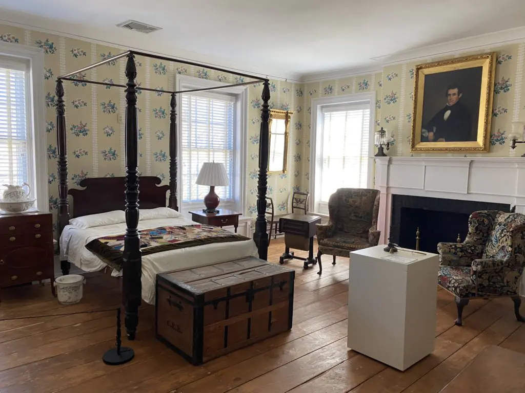 A large wooden bed with canopy support sits in the center of the room. At the foot of the bed is a colorful quilt. In front of the bed is a large wooden chest. There are wooden nightstands on either side of the bed. To the right of the bed is a wooden table and upholstered chair. In front of the chair is a pedestal with objects that can be touched. A painted portrait hangs over the fireplace.