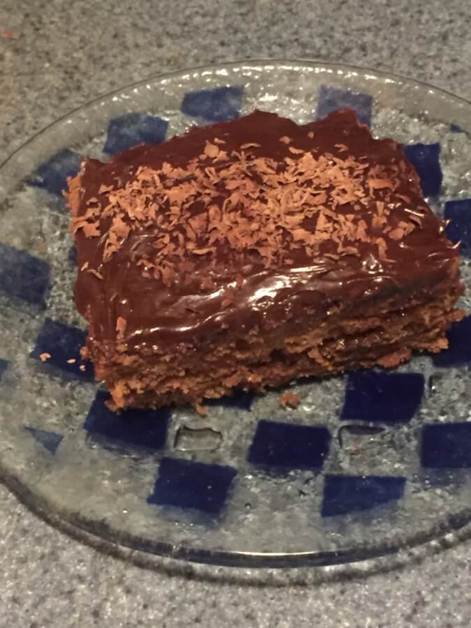 A slice of chocolate cake topped with chocolate icing and chocolate shavings sits on a clear glass plate.