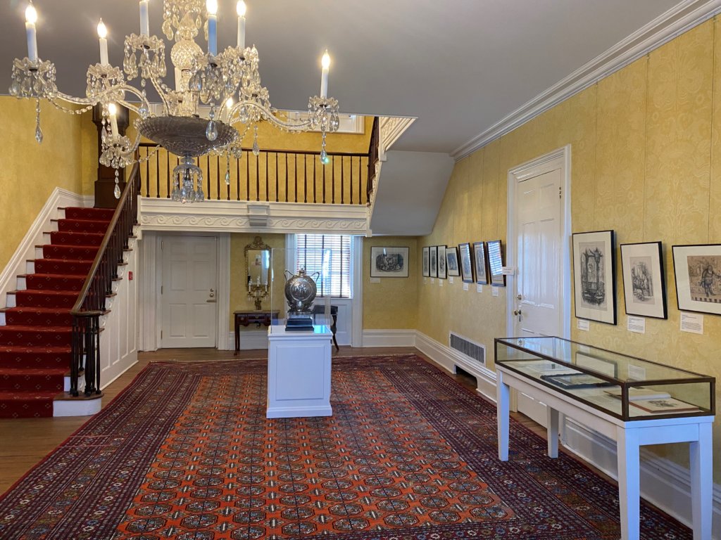 A chandelier hangs in the top left corner. In the back left corner of the room is a grand staircase. The center of the room has a silver canteen on display in an enclosed case. On the right of the image, several framed prints hang on the wall. Below the prints is a glass case with additional artifacts.