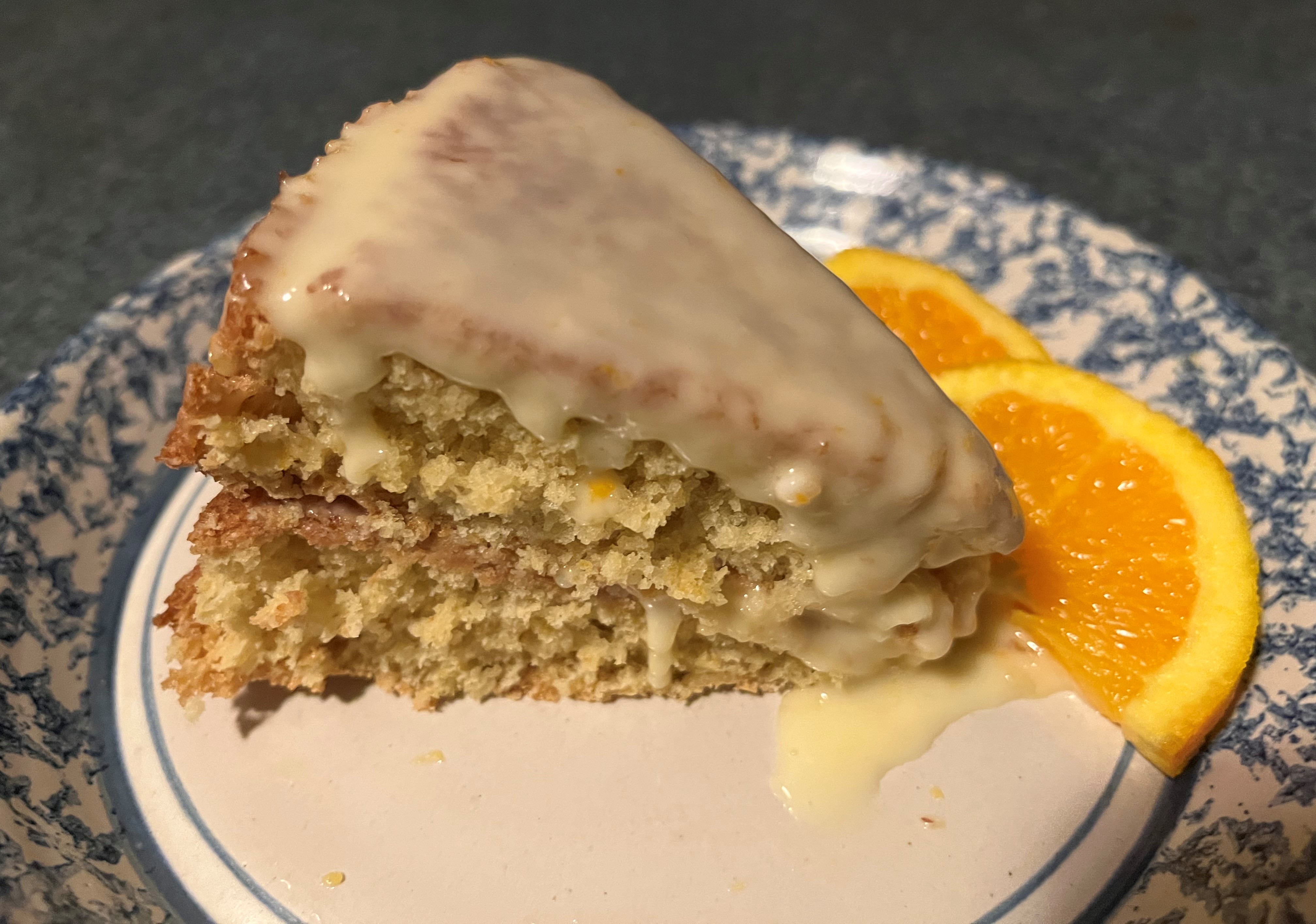 A slice of orange cake topped with icing sits on a plate next to two orange slices.