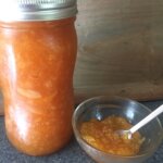 A glass jar full or orange marmalade sits next to a bowl with spoon and orange maramalade.