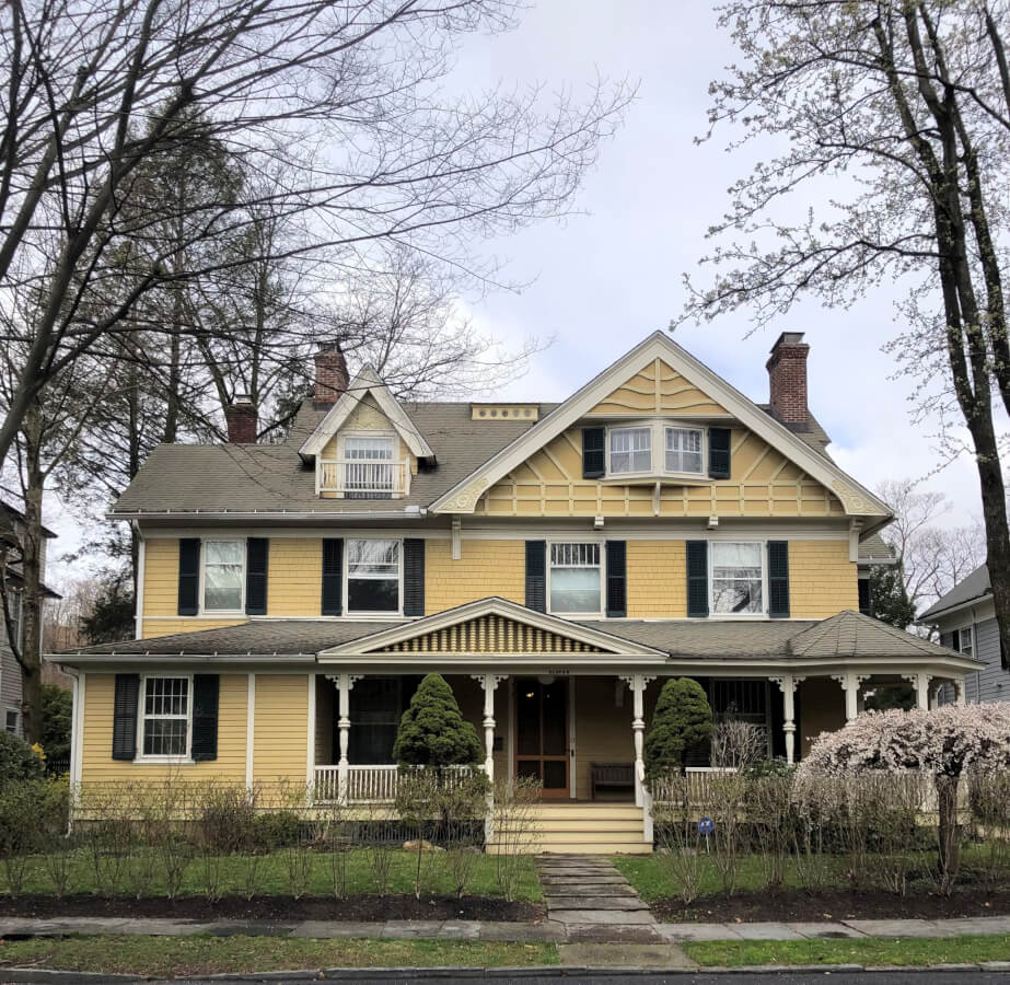 A yellow Queen Anne style home with front porch and green bushes in front.