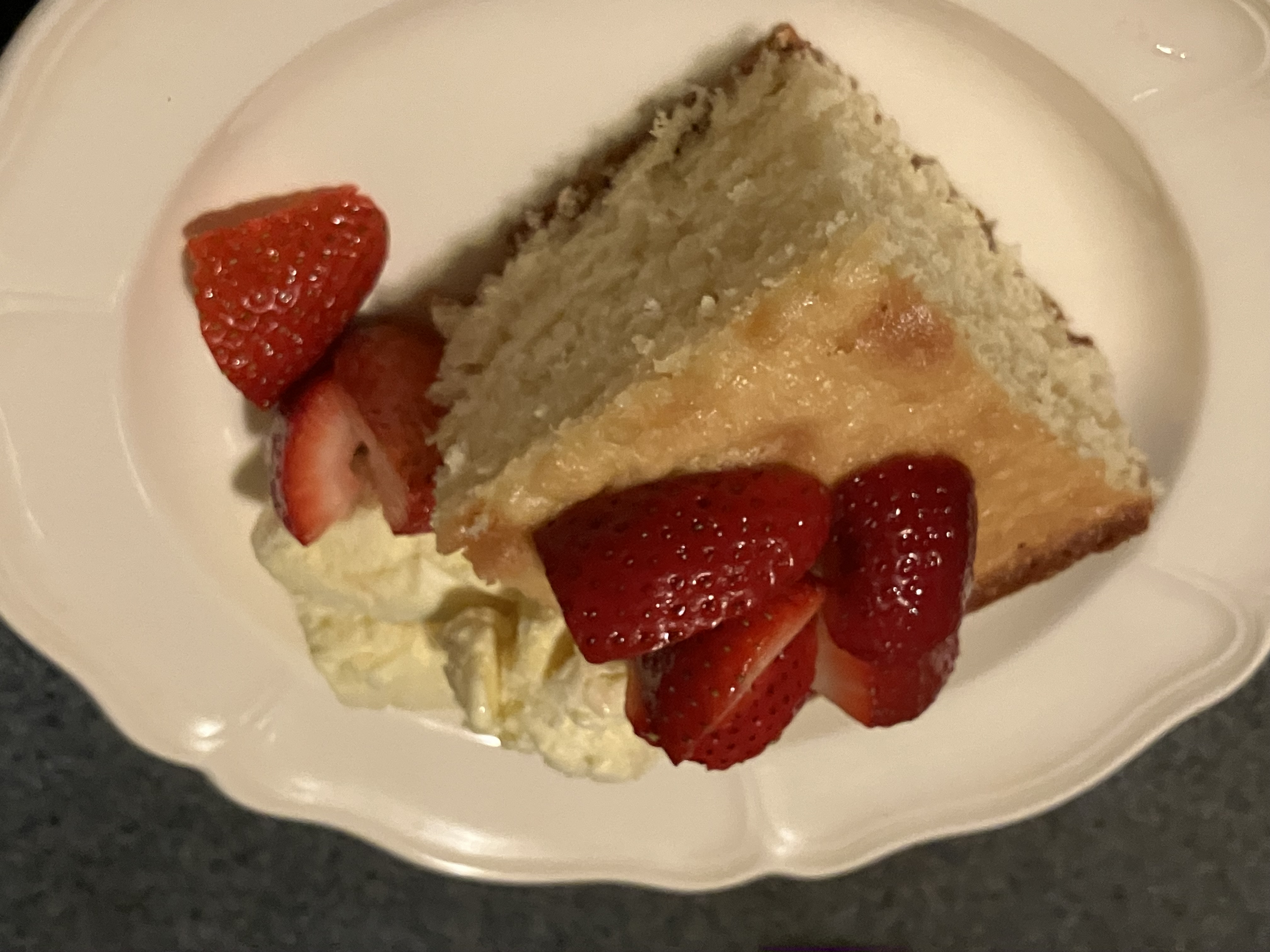 A square sliced of baked cake on a round white dish. The cake is topped with strawberry slices. Whipped cream and strawberry slices are next to the cake.