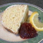 A slice of yellow cake topped with powdered sugar on a plate next to jam and a lemon slice.