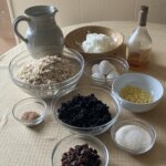 Several wet and dry ingredients laid out in a pitcher, bottle, and various bowls on a table.