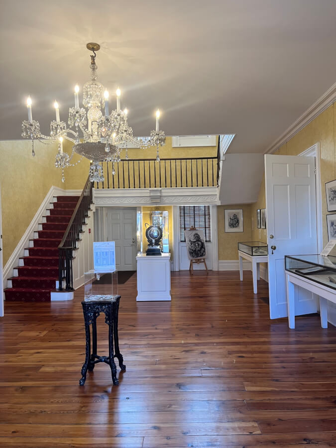 Open room with yellow wallpaper. A grand staircase in the background. A chandelier in the foreground.