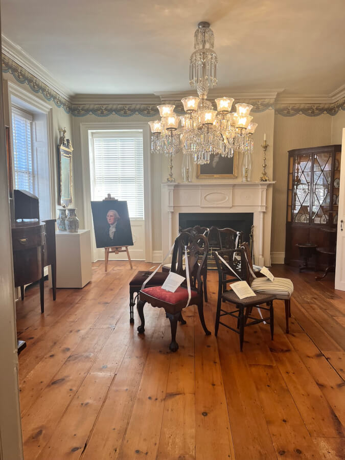 Room with chairs arranged in a circle under a chandelier in the center. A portrait of George Washington stands to the left of the fireplace. A china cabinet is to the right of the fireplace.