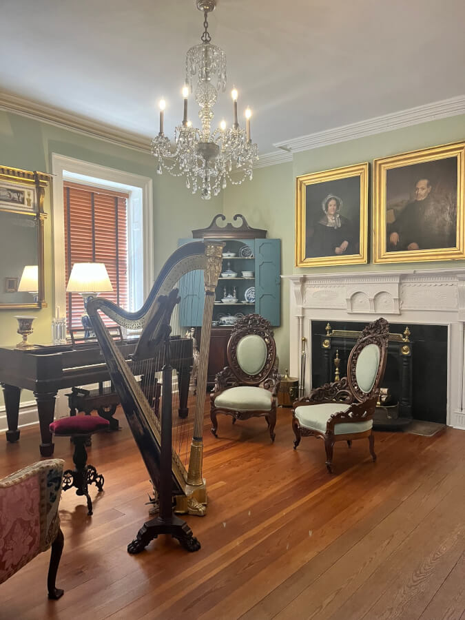 A room with a piano, harp, and music stand. Multiple chairs are arranged around the instruments. Two portraits hang above the fireplace in the background.