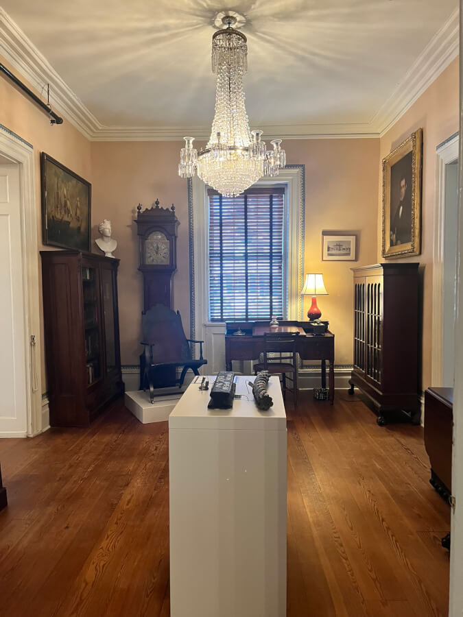 Room with various office furniture including a desk, chairs, bookcases and a grandfather clock. A chandelier hangs from the ceiling. Objects on a white pedestal in the foreground.