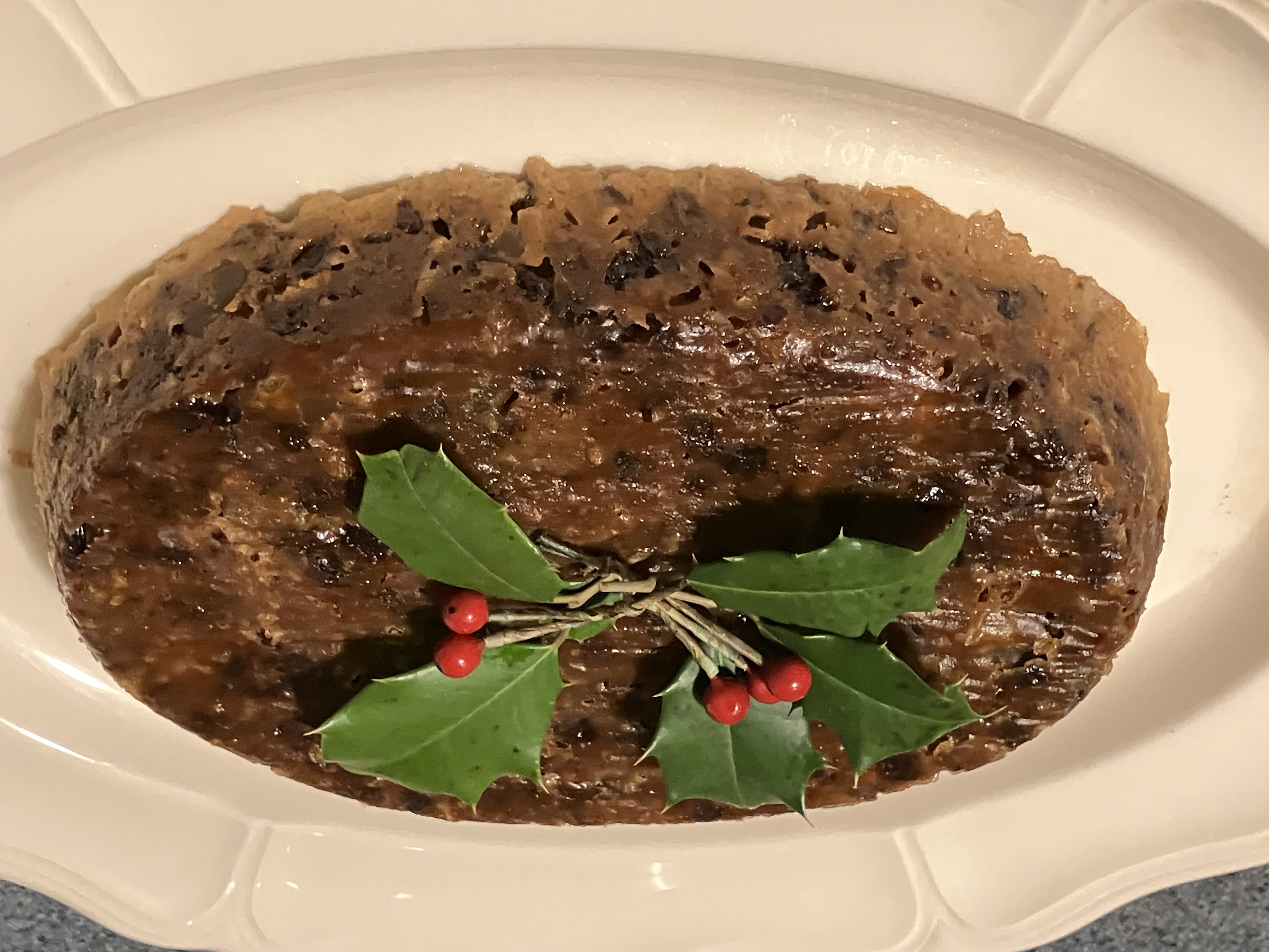 A baked plum pudding on an oval plate with holly leaves and berries on top.