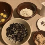 Eggs, flour, dried fruit, and spices arranged in bowls and on plates on a table.