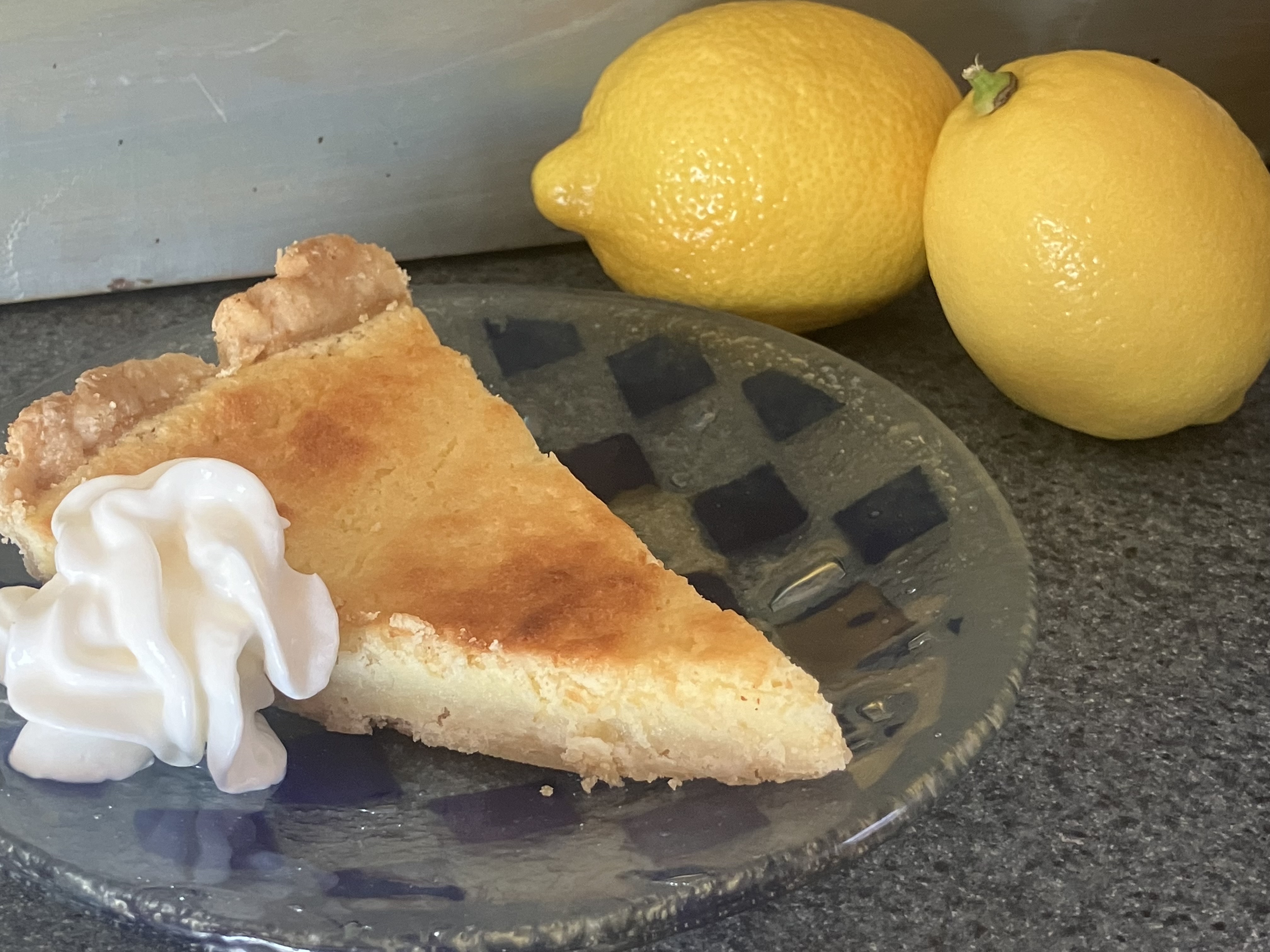 A baked slice of Lemon Pie sits on a plate with whipped cream. Two lemons sit next to the plate.