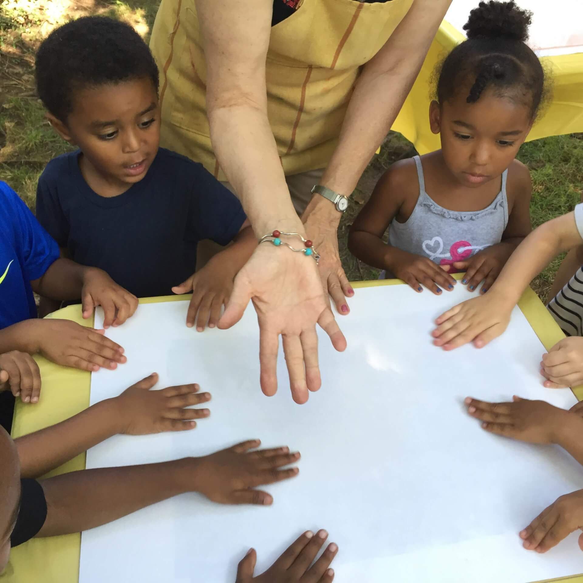 Young children gathered around a table getting ready to make an art project on a large, white piece of paper.
