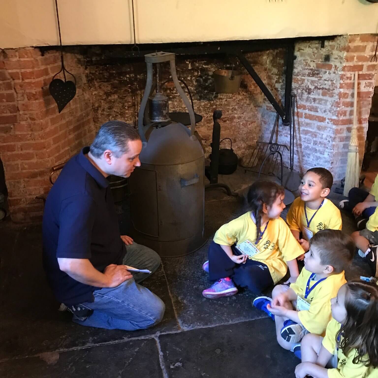 A group of young children wearing yellow t-shirts seated on the hearth in front of the antique fireplace. They are talking with a male curator.